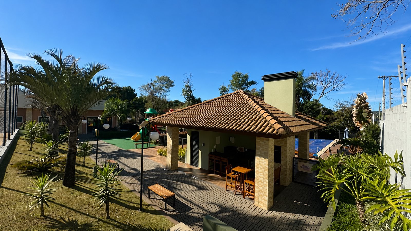 Espaço gourmet coberto do condomínio, com churrasqueira, bar e área de estar ao lado do playground